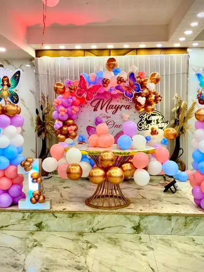 The main stage for a butterfly-themed first birthday party. The setup features a custom backdrop, a circular balloon arrangement in pastel and chrome colors, and fairy character cutouts on pillars.