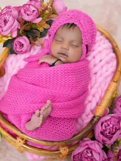 A newborn is wrapped in a bright pink knit swaddle and matching bonnet, nestled in a basket surrounded by flowers. This cozy and colorful setup is perfect for a sweet and classic baby portrait.