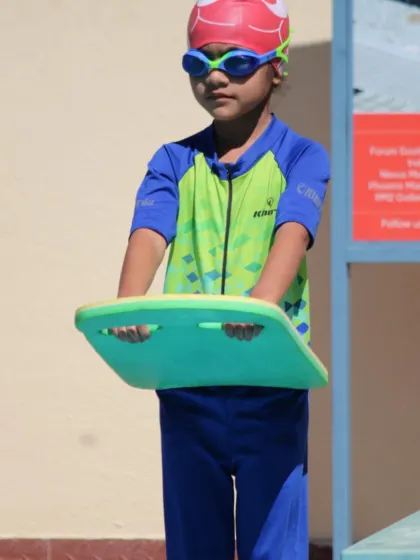 A young swimmer stands ready with her kickboard. This tool is essential in our beginner classes for helping students develop a strong, effective kick while maintaining buoyancy.