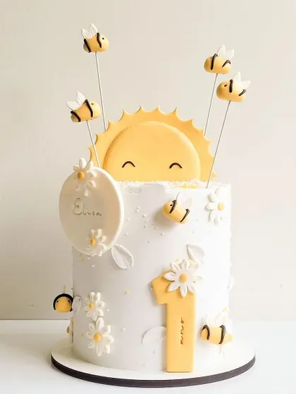 A "You are my sunshine" themed cake for a first birthday. This cheerful cake is decorated with a smiling sun, buzzing bees, and daisies.