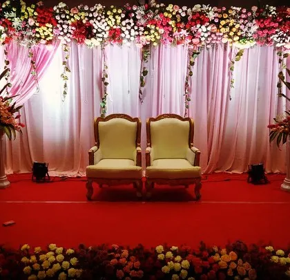 This is a classic and regal stage design for a wedding reception. The setup includes two elegant throne-like chairs for the couple, set against a backdrop of pink drapes and a lush floral border.