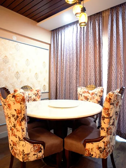 A formal dining area with custom upholstered chairs in a floral pattern. The space is framed by heavy curtains and features a dark wood ceiling panel with pendant lights.