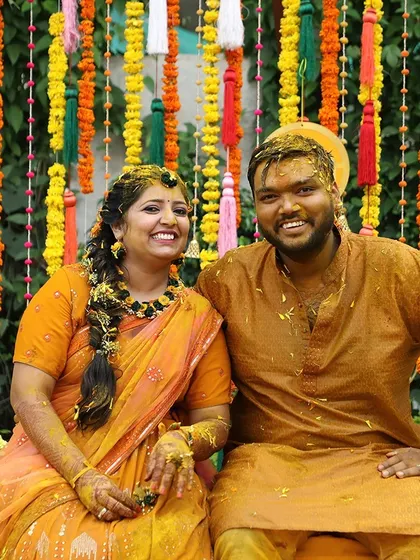 A happy couple posing during their Haldi ceremony, with a backdrop of colorful hanging tassels.