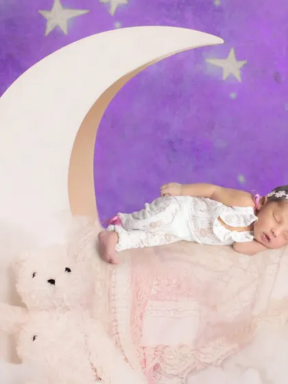A sweet newborn portrait on a moon prop against a starry purple night sky backdrop.