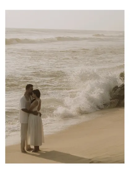 A wide shot of the couple embracing on a sandy beach, with waves crashing against the rocks nearby. The image captures both the power of the ocean and the quiet strength of their connection.