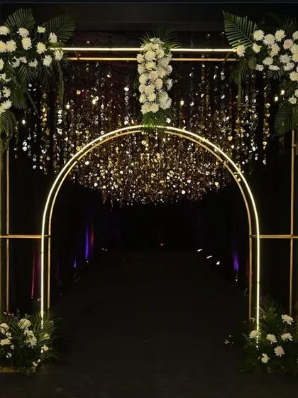 A sleek and modern entrance arch for a black-themed event. The minimalist gold frame is illuminated and decorated with white flowers and hanging crystals.