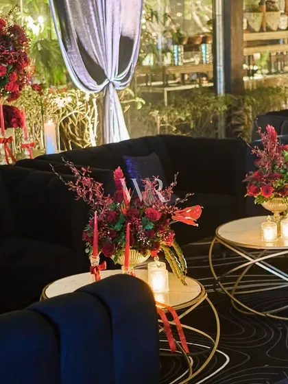 A lounge area with black velvet sofas and a swirling patterned rug. The space is made intimate with low coffee tables, each adorned with red floral arrangements and candles.