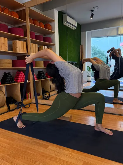 A student in a deep back-bending lunge, using a strap to connect her hand to her foot. The mirror reflects her beautiful alignment.