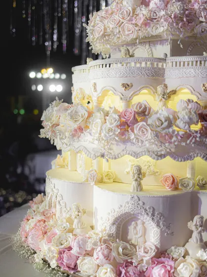 The lower tiers of this grand wedding cake showcase the detailed architectural elements, including arched windows, delicate balustrades, and clusters of handmade sugar roses, all softly lit from within.
