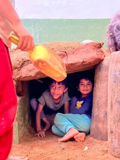 Two boys peek out from inside their newly constructed cobb house. They have created their own private, cozy space, a perfect nook for stories and secrets.