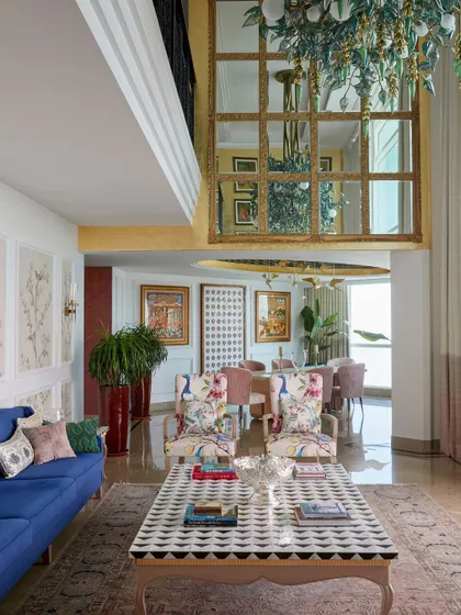 This view shows the connection between the living and dining areas, with the mezzanine level overlooking the space. A large mirror with a gold frame reflects the chandelier, adding to the sense of grandeur.