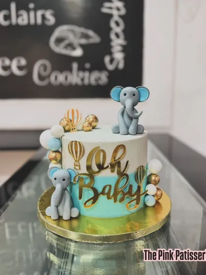 An "Oh Baby" shower cake with an adorable elephant theme. The cake features a blue and white watercolor effect, gold hot air balloon details, and two cute fondant elephants.