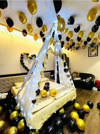For a stylish and modern romantic look, I use black and gold balloons. This room decoration includes a canopy with fairy lights and balloons scattered on the floor and bed.