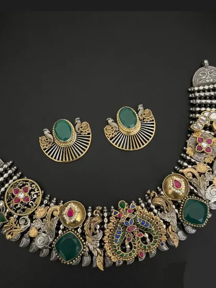 An intricate masterpiece from our fusion collection. This necklace set combines an oxidised silver base with gold-plated elements, green stones, and detailed peacock and floral motifs for a truly one-of-a-kind look.