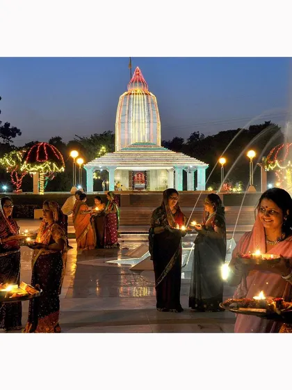 Devotees hold diyas during an evening ceremony outside an illuminated temple. The warm light from the lamps and the joyful expressions capture the beautiful atmosphere of a community united in faith.