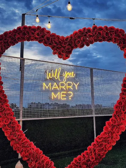 A close-up shot of the iconic red rose heart arch against a cloudy evening sky. The "Will you MARRY ME?" neon sign glows from within, highlighting the detail and craftsmanship of this classic proposal element.