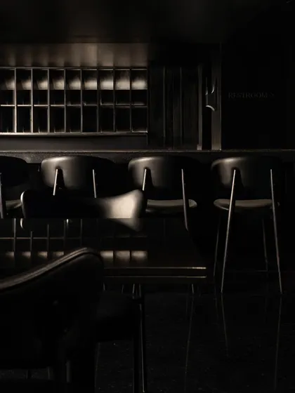 The bar area at Lair, a composition of shadow and minimal light. The design uses focused lighting to highlight the bottles and the dark, fluted bar front, creating a dramatic focal point.