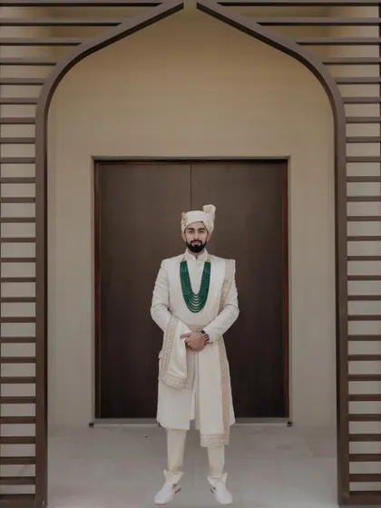 A regal portrait of the groom in his ivory sherwani, complemented by a stunning emerald necklace. The architectural arch frames him perfectly, adding to the grandeur.