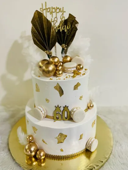 A grand two-tiered white and gold cake for a 60th birthday. This luxurious design is adorned with gold leaf, macarons, and dramatic fan toppers.