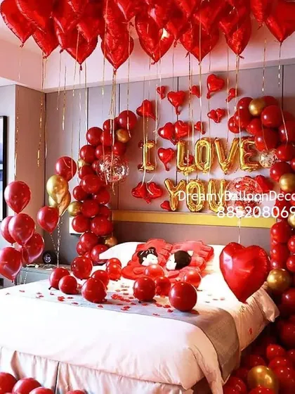 A luxurious red and gold themed romantic room decoration. The bed is framed by a balloon arch, with 'I LOVE YOU' spelled out in foil balloons and heart-shaped balloons filling the ceiling.