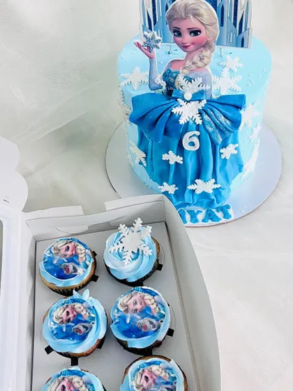 A beautiful Frozen-themed cake featuring Queen Elsa. The design includes a striking blue dress made of fondant and delicate snowflake details, with matching cupcakes ready for the party.