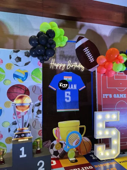 A personalized sports jersey with the birthday boy's name and age, a lit-up number '5', and various sports equipment props make this corner of the party extra special.