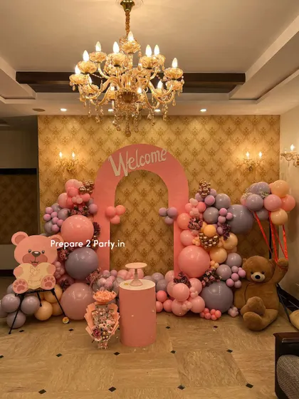 A wide shot of a lavish welcome home decoration set under a beautiful chandelier. The pink arch is flanked by elaborate balloon garlands and cute teddy bear props, creating a grand and festive atmosphere.