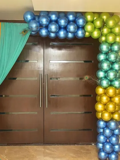 This entrance decor for a Krishna-themed event uses drapes and balloons in colors associated with the theme, like peacock blue and green. A welcome board with a Krishna graphic completes the look.