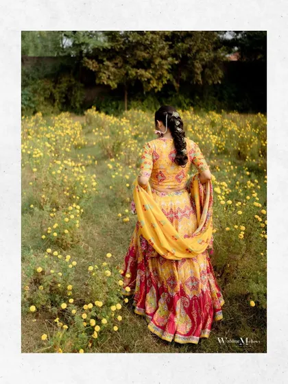 The bride walks through a field of yellow flowers, her colorful lehenga creating a beautiful contrast. This is a dreamy and romantic portrait.