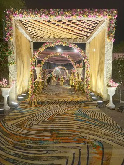 A beautiful evening entrance featuring a wooden trellis roof and floral arches leading into the venue. The combination of a uniquely patterned carpet and warm lighting creates a welcoming and stylish entryway.