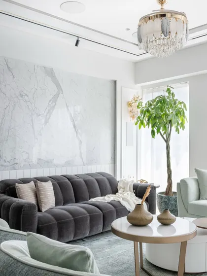 A close-up of a luxury living room where a plush tufted charcoal sofa adds depth against a white marble wall. A statement chandelier and track lighting provide layered illumination, while a large indoor plant brings freshness and vibrancy.