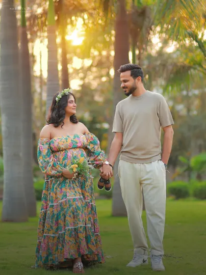A romantic outdoor portrait of a couple holding hands and baby shoes. The golden light and floral gown create a warm and dreamy atmosphere.