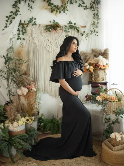 A full-length portrait of a mother-to-be in a classic black off-shoulder gown. The contrast of the dark dress against the light, airy boho backdrop is stunning.