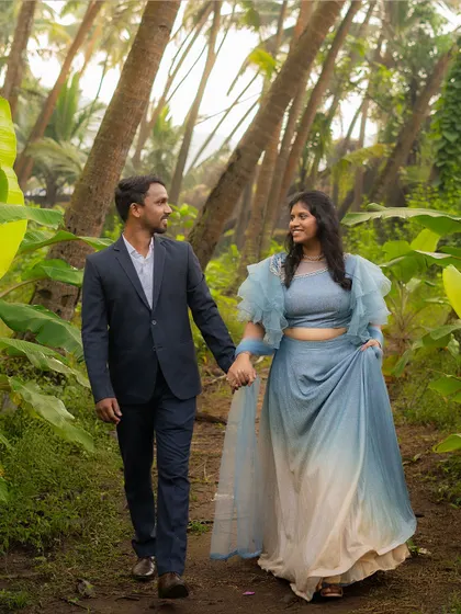 A couple dressed in elegant blue attire walks hand-in-hand through a tropical path, the vibrant green leaves creating a beautiful natural frame for their pre-wedding photo.