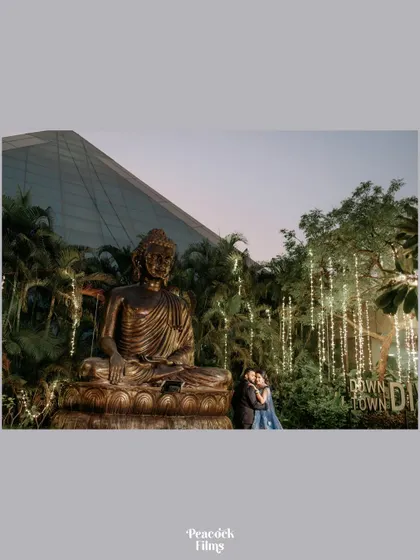 A wide, atmospheric shot of the couple posing in front of a large Buddha statue and beautifully lit trees, capturing the serene and grand ambiance of their event.