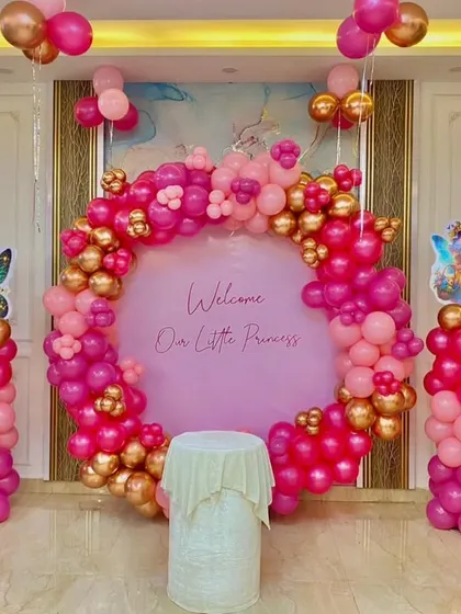 A gorgeous "Welcome Our Little Princess" setup. The circular backdrop is framed by a rich pink and gold balloon arch, with fairy cutouts on matching balloon pillars, creating an elegant and celebratory look.