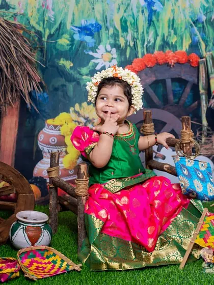 A charming photo of a toddler in a traditional pavadai, sitting in a rustic village-themed set. Her sweet, shy expression and the colorful props create a delightful scene.