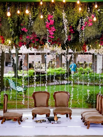 This enchanting mandap design for a garden wedding features a ceiling of lush greenery and pink flowers, with hanging pearl strings that add a touch of fairytale magic.