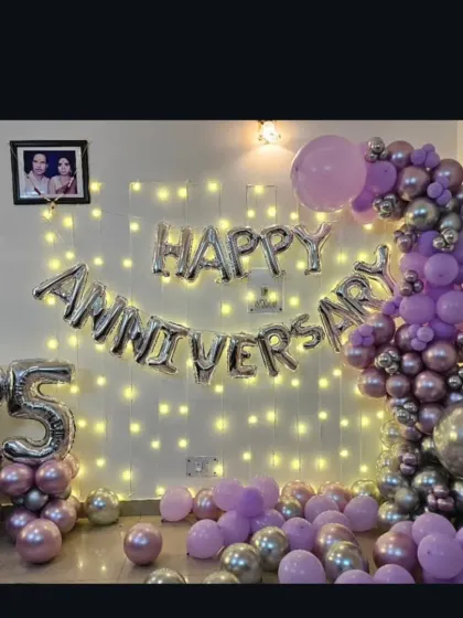 A beautiful 25th-anniversary decoration at home with a fairy light wall, 'Happy Anniversary' foil balloons, and a purple and silver balloon garland.