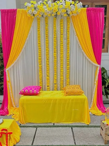 A clean and elegant Haldi stage setup with bright pink and yellow drapes. The backdrop is accented with vertical strings of white flowers, creating a simple yet beautiful space for the ceremony at home.