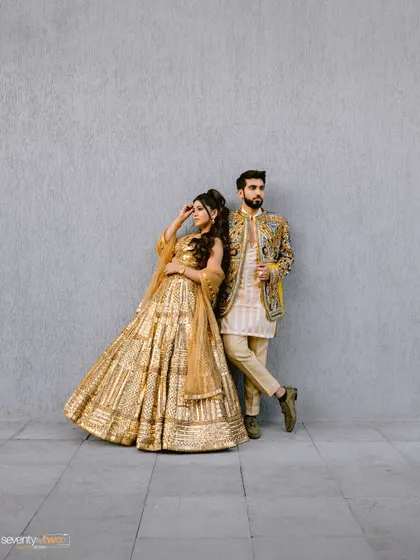 A full-length, editorial-style portrait of the couple posing against a simple grey wall, letting their stunning outfits take center stage.