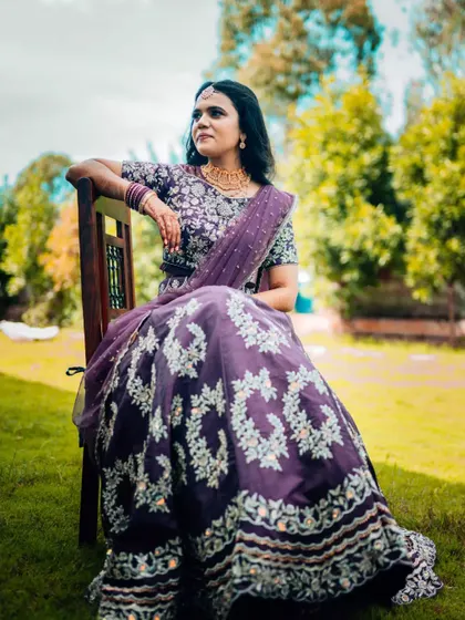 A full-length portrait of a woman in an elegant purple lehenga, seated outdoors. The natural light and green background make the colors of her outfit pop.
