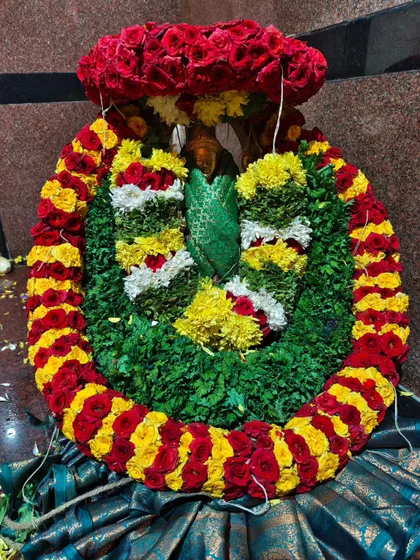 A close-up of a deity's crown and face, adorned with a large, intricate garland made of red roses, yellow flowers, and green leaves.