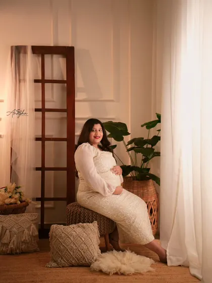 A bright and airy studio portrait. The mother-to-be is glowing in a beautiful white lace dress, surrounded by our light-filled boho setup.