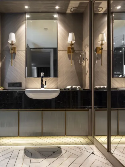 A bathroom in the Jor Bagh Residence, showcasing a sophisticated material palette. A custom vanity with a black marble countertop is set against a wall of diagonally laid textured stone, with elegant brass-finished fixtures and sconce lighting.