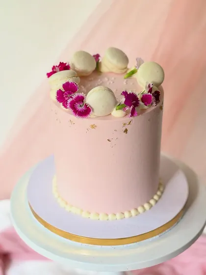 A chic and simple pink cake with a subtle gold leaf accent. The top is elegantly decorated with a ring of white macarons and fresh pink carnations.