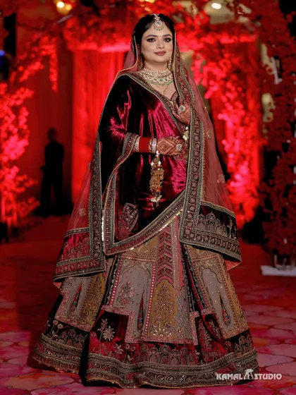 A full-length portrait of the bride in her majestic velvet lehenga, against a backdrop of red flowers.