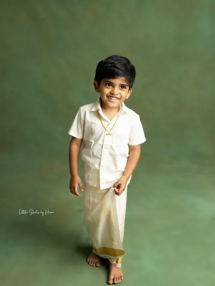 A handsome young boy in a traditional veshti, standing proudly for his portrait. The simple green background makes his bright smile pop.
