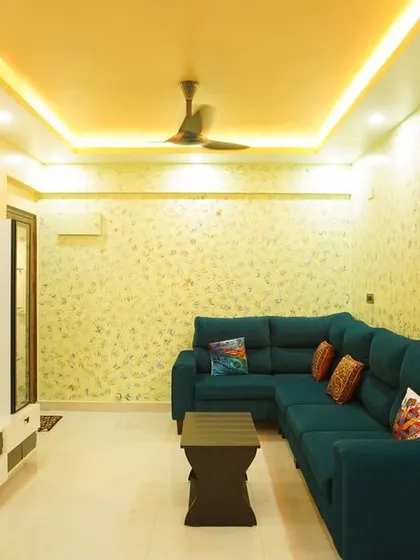 A comfortable living room with a large teal sectional sofa and textured wallpaper. The false ceiling with integrated lighting adds a modern touch to the cozy space.