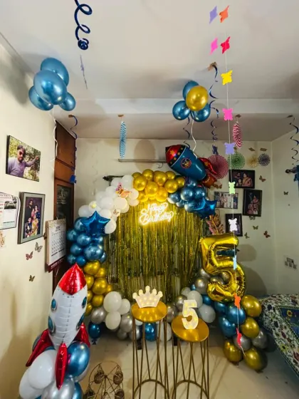 For this 5th birthday, I designed an outer space theme for a celebration at home. The decoration features a large balloon arrangement with rocket ship foil balloons, a gold fringe backdrop, and a rented 'Happy Birthday' neon sign. The large foil number '5' makes the milestone clear.